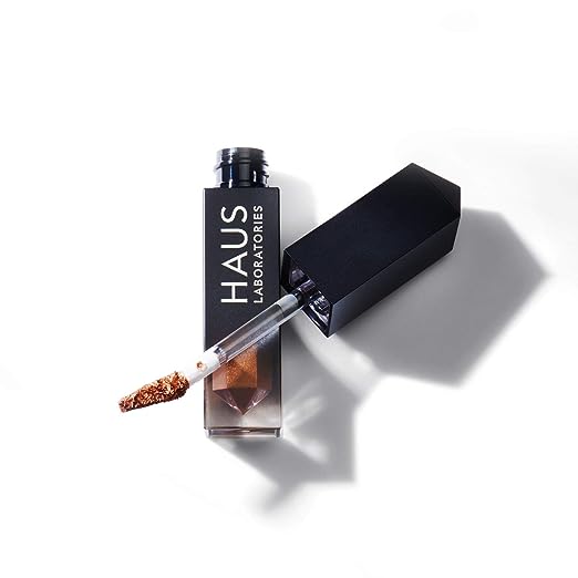 An image of a bottle of Haus Labs liquid eyeshadow. The bottle is open, with the cap and built-in brush resting on top of the bottom of the bottle, which is lying flat against a white background. The eyeshadow is a golden brown color. The bottle reads "Haus Laboratories".
