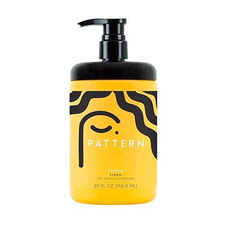 An image of a bottle of Pattern brand styling cream. The bottle is black with a yellow label and black pump top. The label has a stylized illustration of a woman's face and curly hair. The label reads "Pattern Styling Cream."